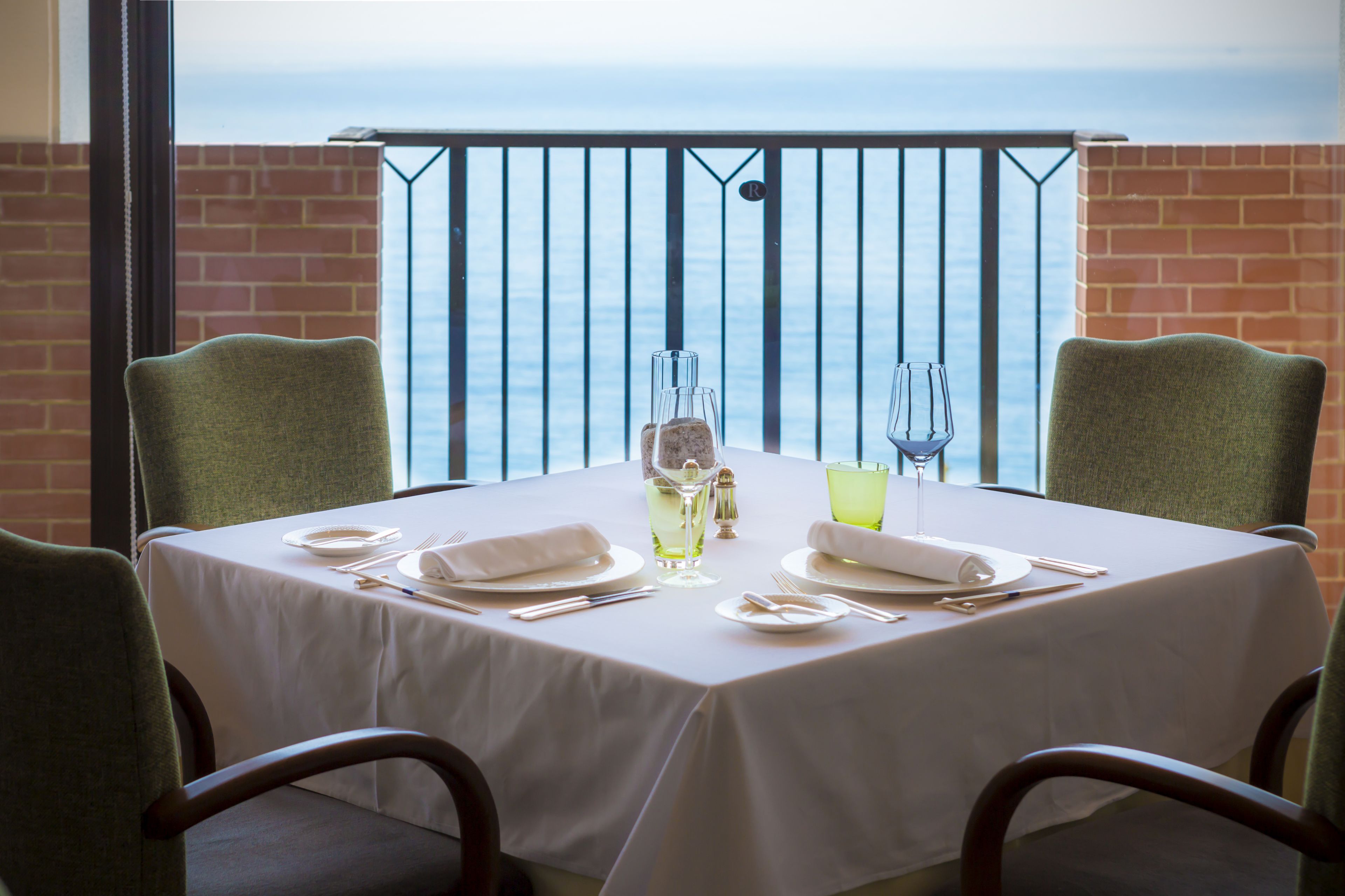 A dining table set with white tablecloth, plates, cutlery, and glasses, positioned on a balcony with a view of the sea.