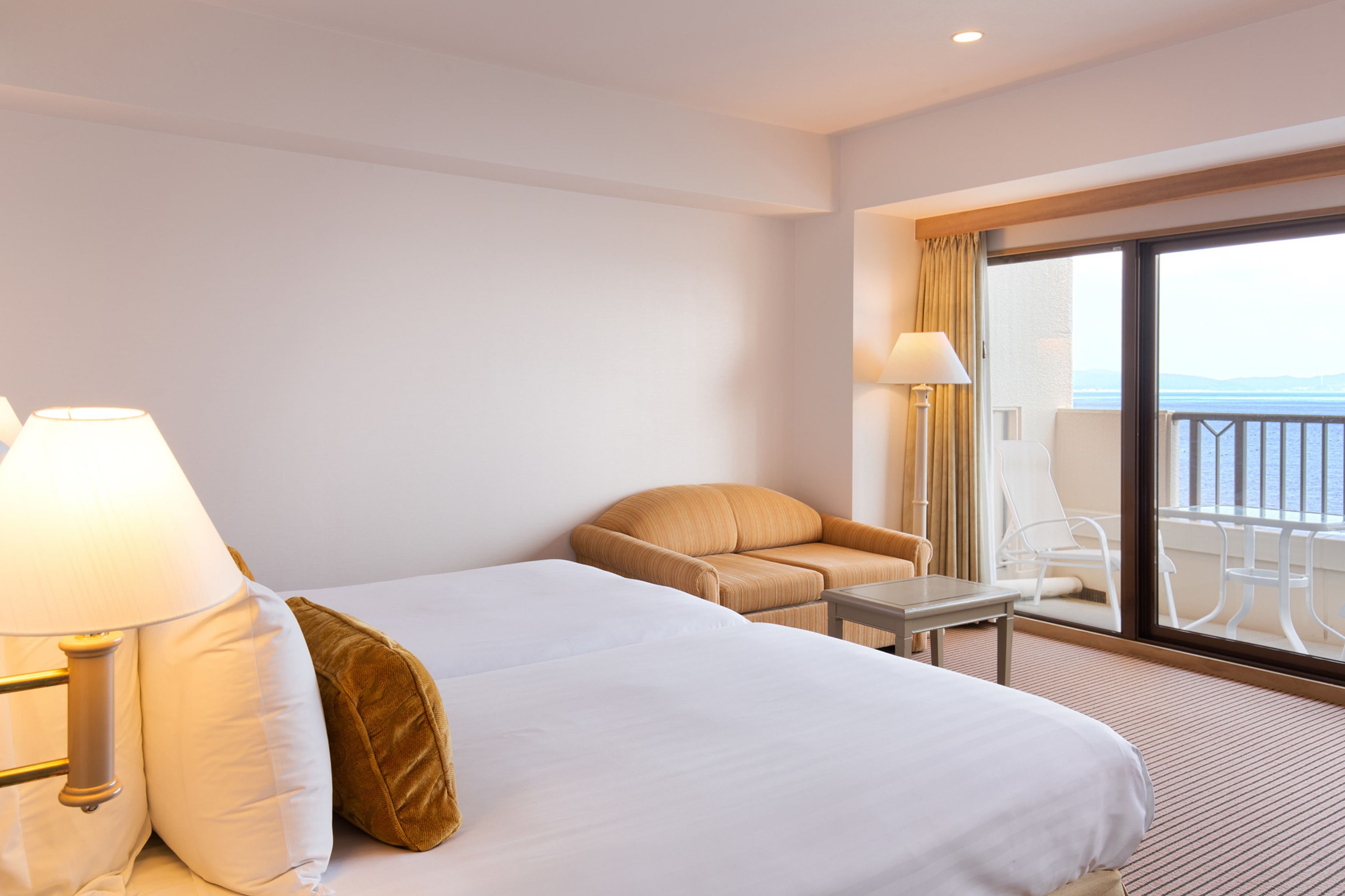 A hotel room with two beds, a small table, a chair, and a balcony with a view of the sea.