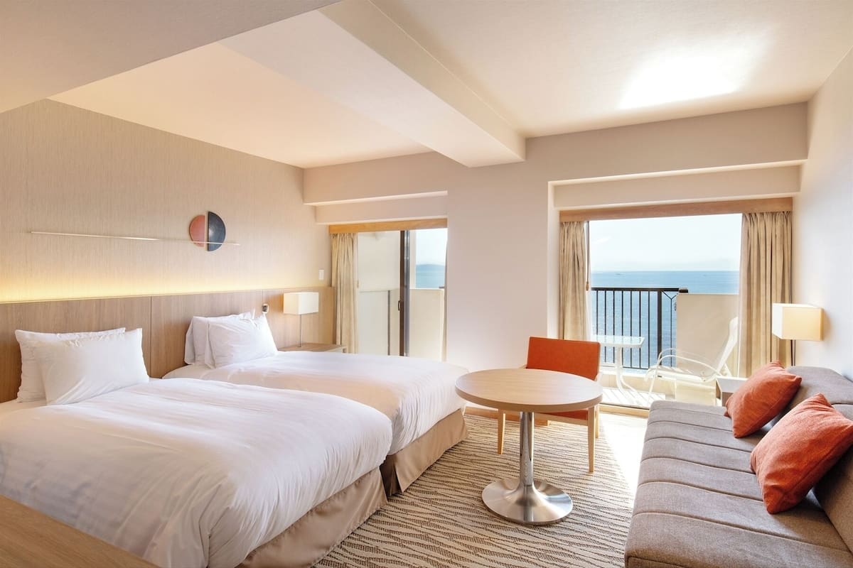 A hotel room with two beds, a small round table, a sofa, and a balcony with a view of the sea.