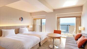 A hotel room with two beds, a small round table, a sofa, and a balcony with a view of the sea.