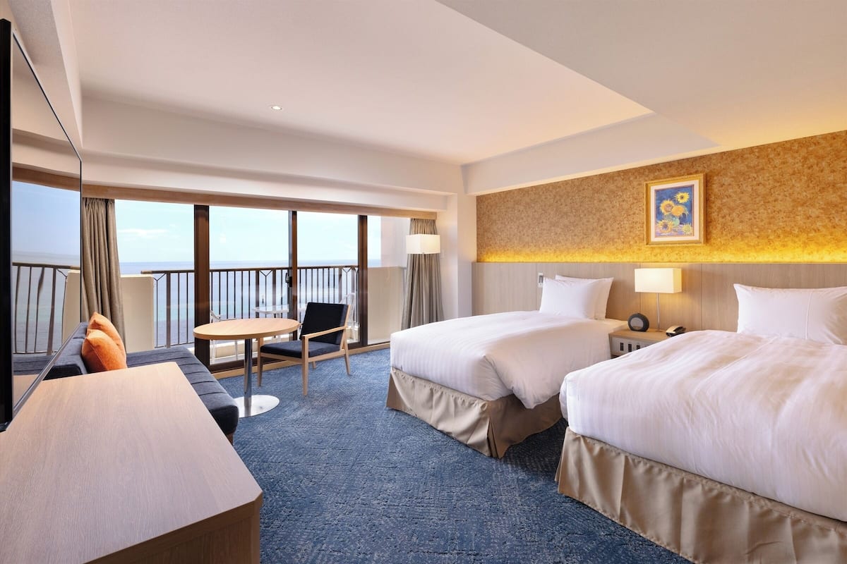 A hotel room with two beds, a desk, a chair, a balcony with a view, and a painting on the wall.