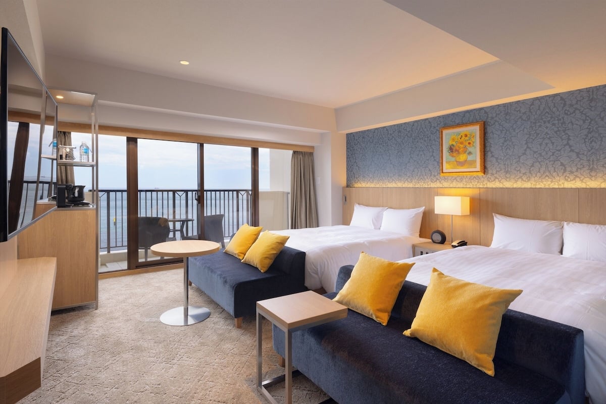 A hotel room with two beds, a sofa, a small table, a TV, and a balcony with a view.