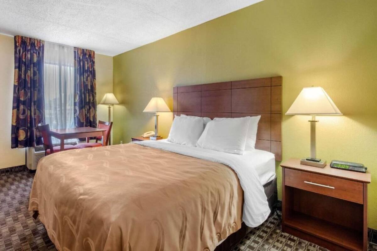 A hotel room with a bed, two bedside lamps, a nightstand with a clock, a chair, a table, and a window with curtains.