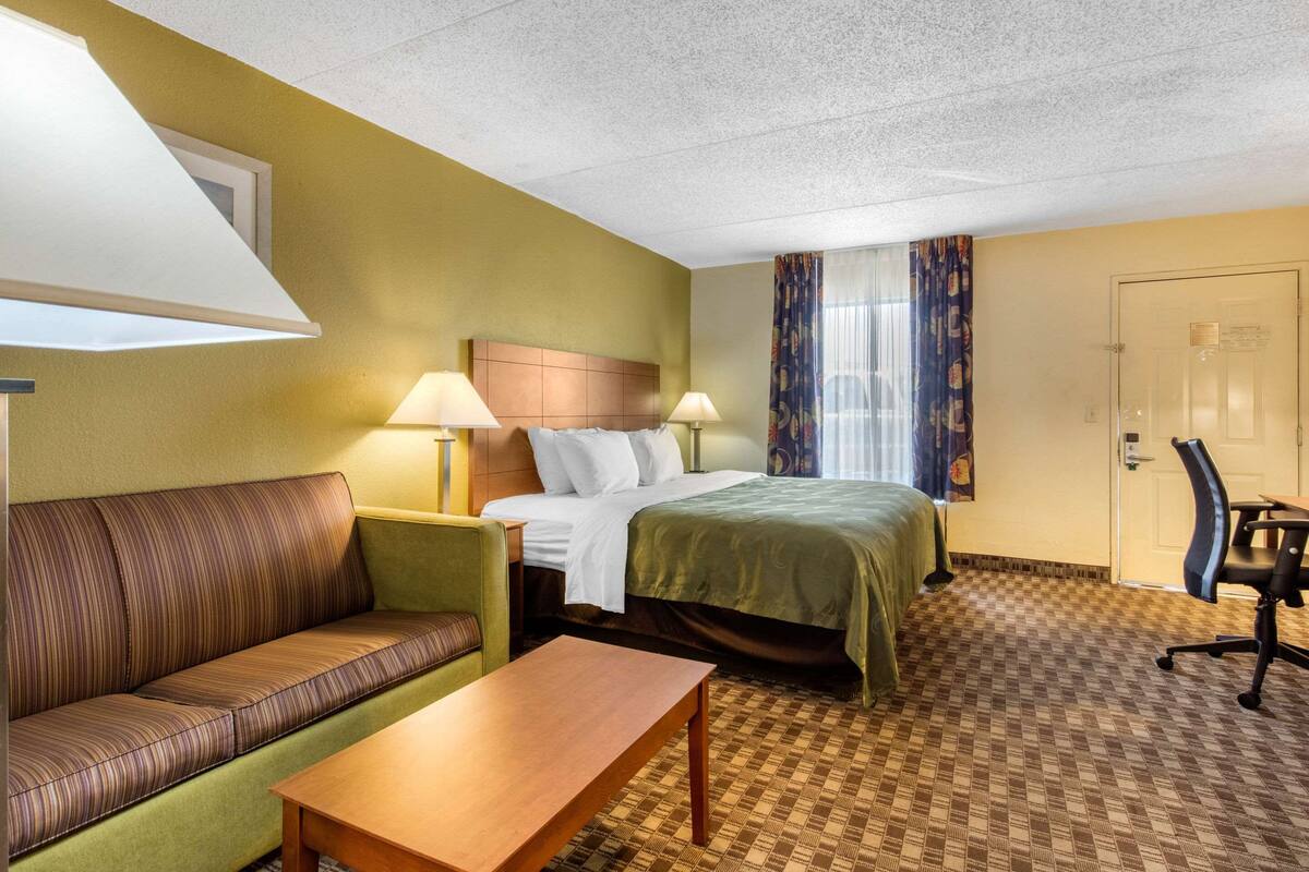 A hotel room with a bed, a green sofa, a wooden table, and a desk with a chair.