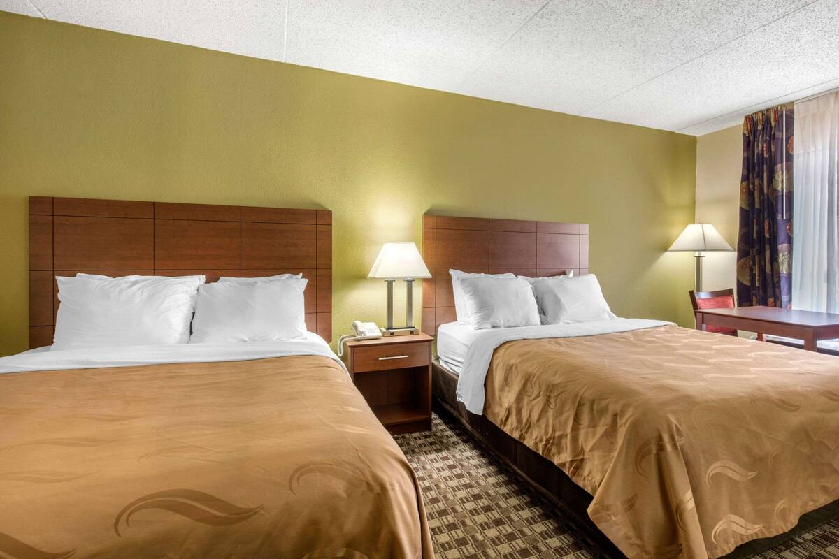 A hotel room with two beds, a nightstand with a lamp, a telephone, and a small table with a chair.