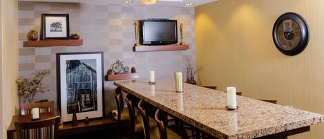 A modern kitchen with a granite countertop, wooden chairs, a television above the counter, and a wall clock.