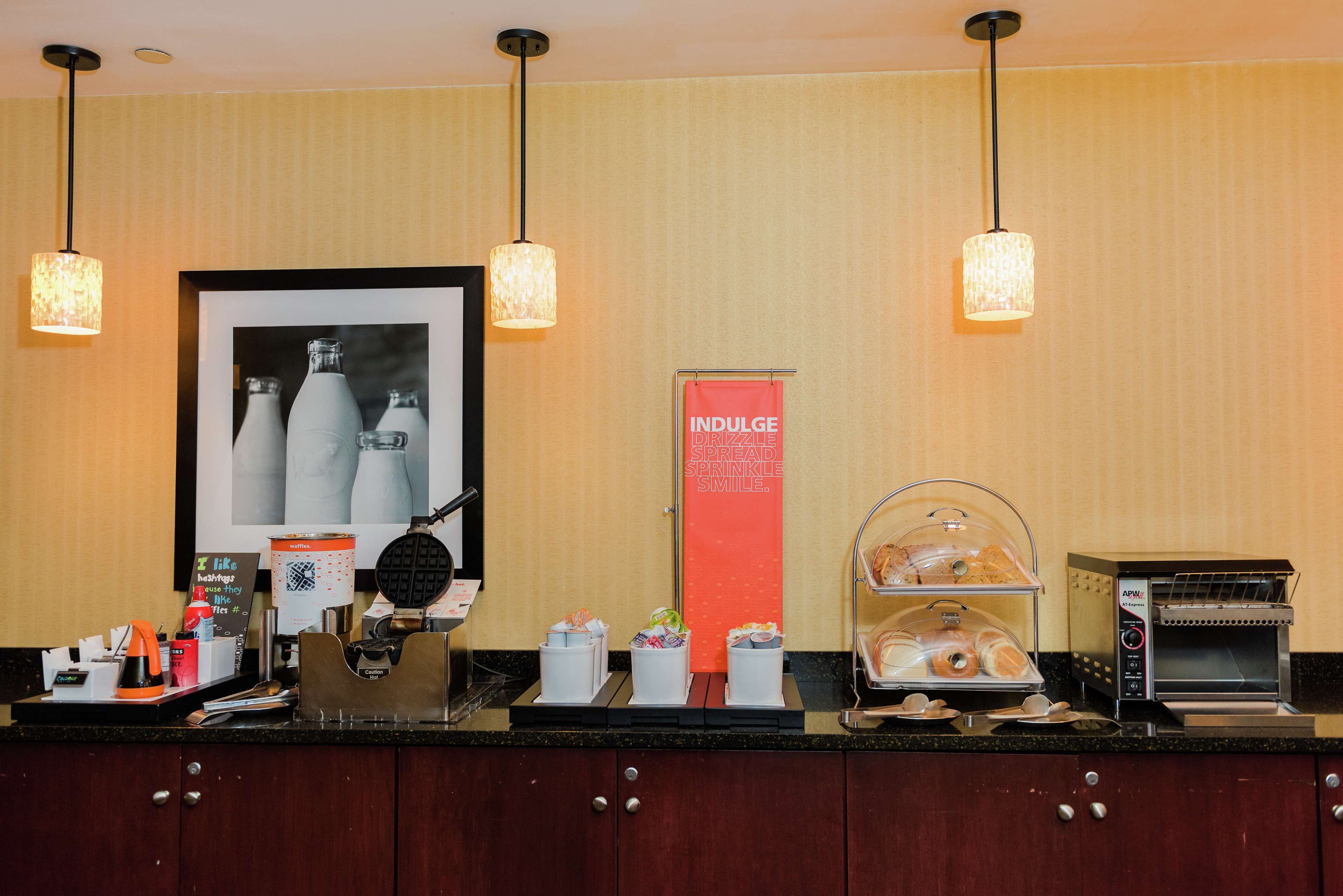 Un espace de petit-déjeuner d’hôtel avec une machine à café, un présentoir de sandwichs et un panneau sur lequel on peut lire : « Savourez, dégustez, partagez, souriez. »