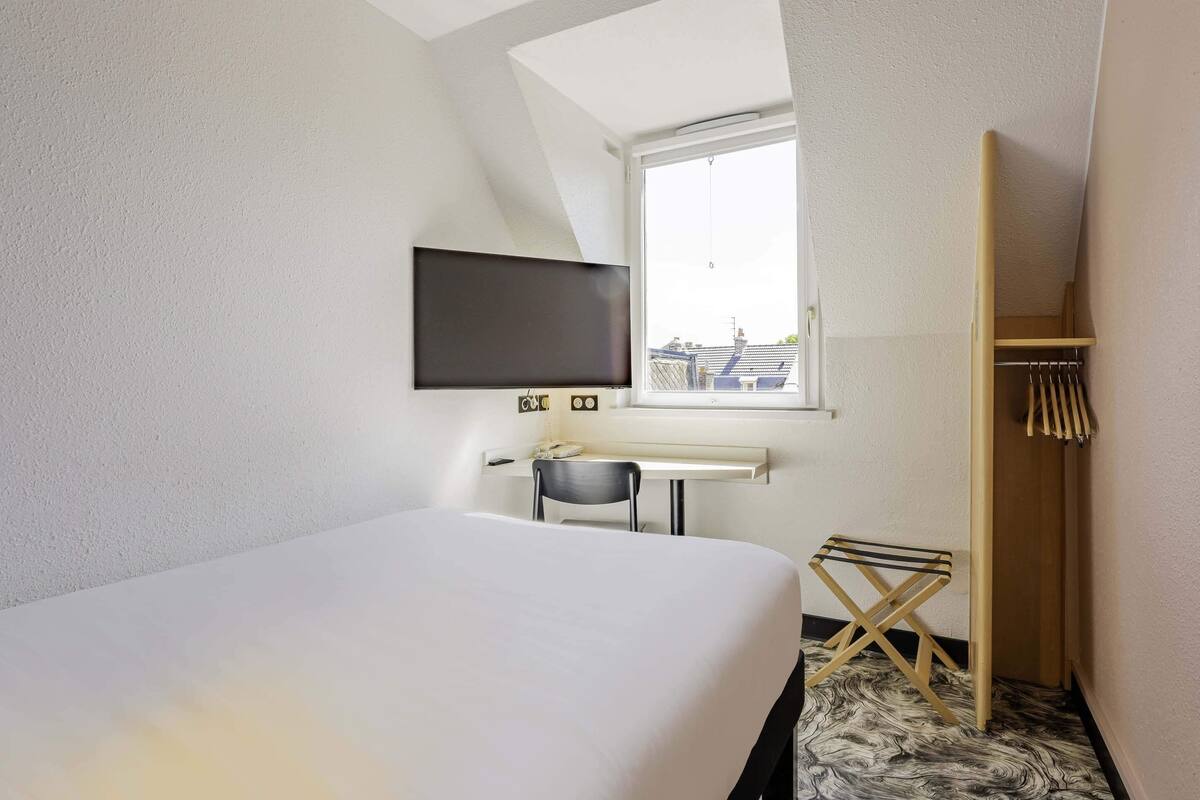 Un dormitorio con cama, escritorio, silla, televisor montado en la pared y ventana con vista a edificios.