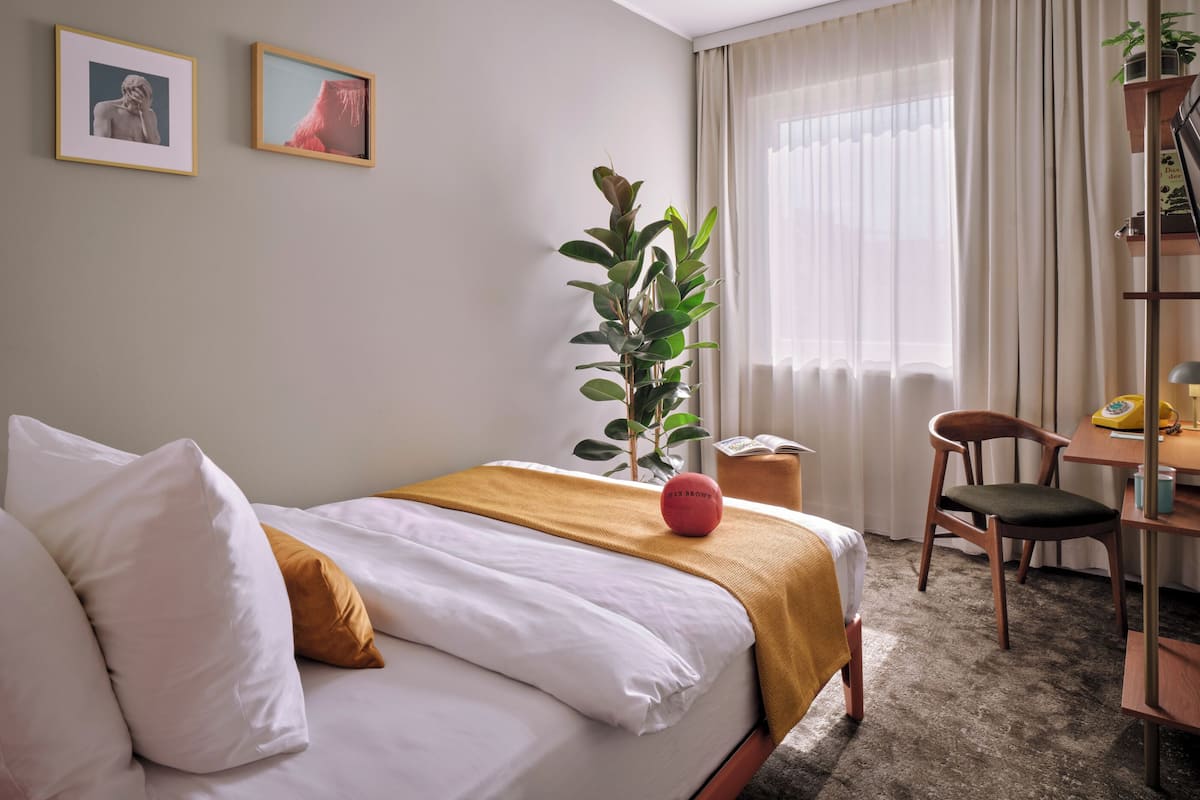 Une chambre d’hôtel bien rangée, avec un lit, un bureau, une chaise, une plante en pot et deux tableaux encadrés accrochés au mur.