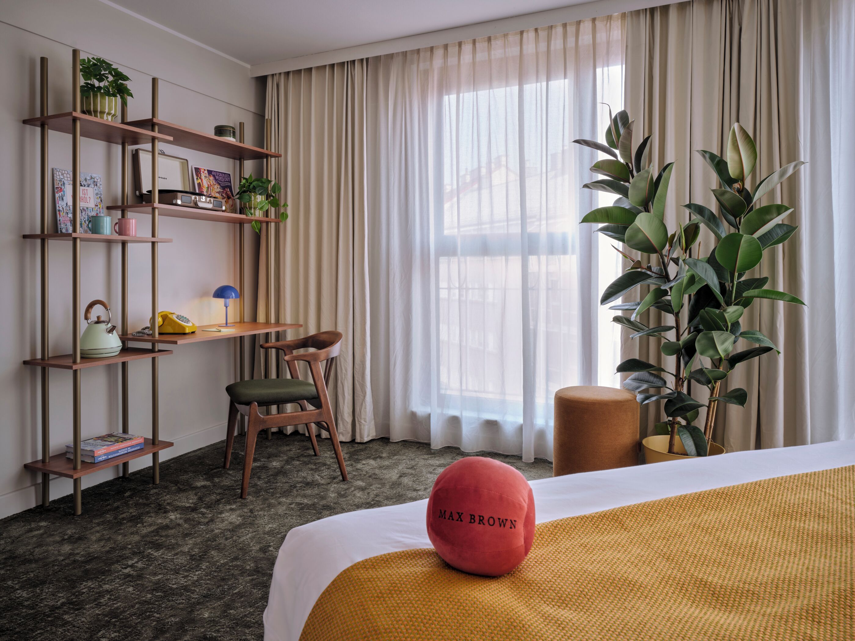A hotel room with a bed, a desk, a chair, a bookshelf, a potted plant, and a window with curtains.