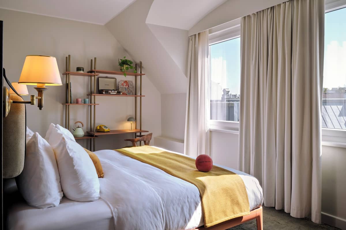 Une chambre d’hôtel avec un lit, un bureau, une lampe, une étagère avec des livres et une plante, et une fenêtre avec des rideaux.