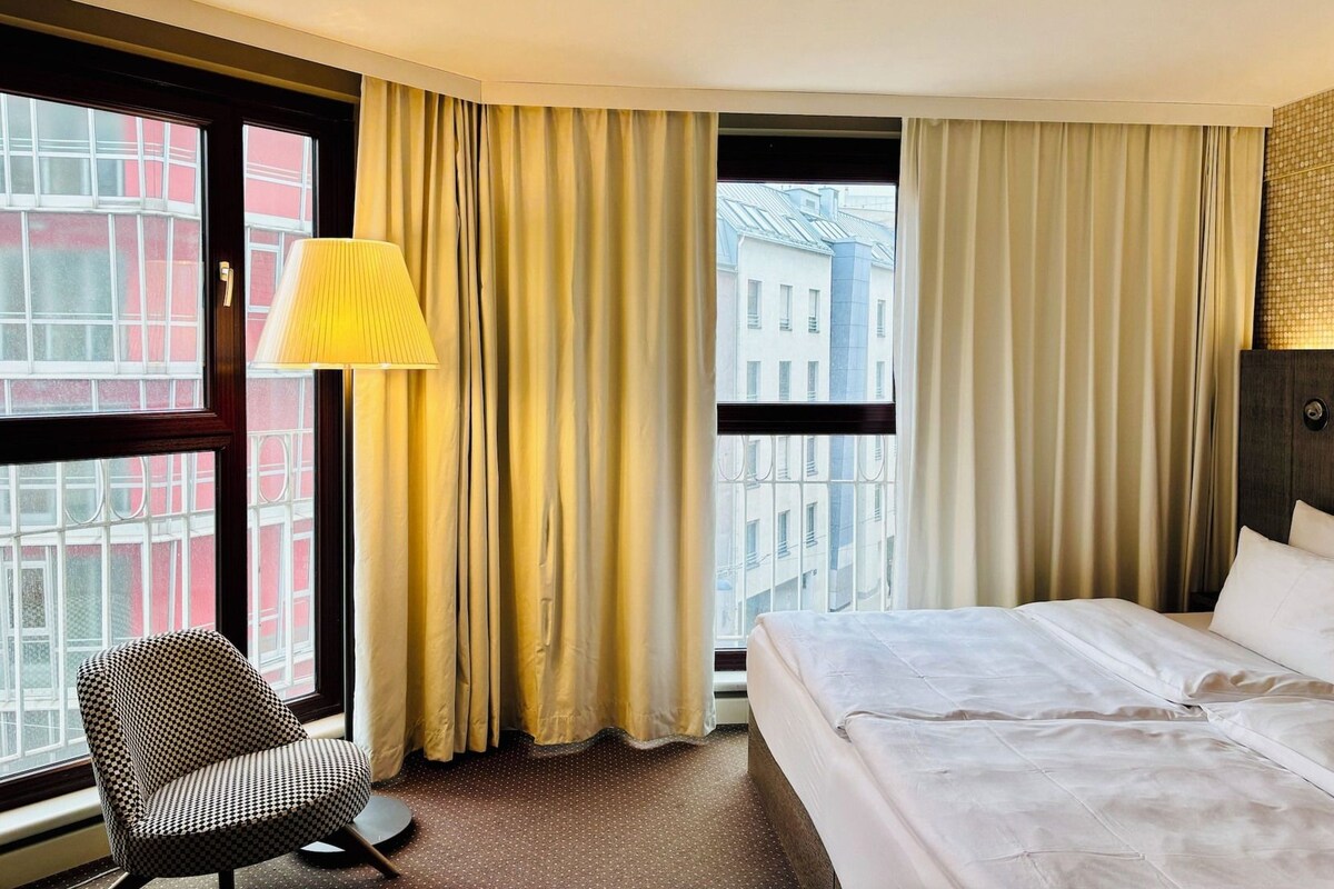 A hotel room with a bed, a chair, a lamp, and a view of buildings through the window.