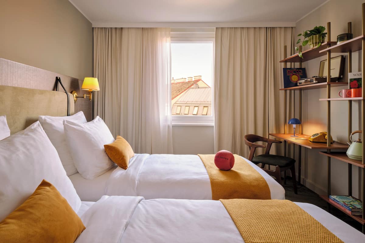 Une chambre d’hôtel avec deux lits, un bureau avec une chaise, une bibliothèque et une fenêtre avec des rideaux.