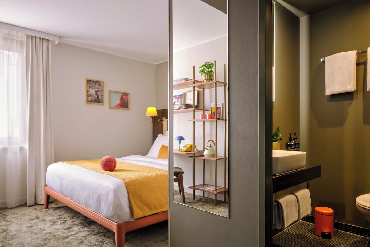 Une chambre d’hôtel comprenant un lit, une table de chevet, un miroir, une étagère avec des articles de toilette et une salle de bain avec un lavabo et des toilettes.
