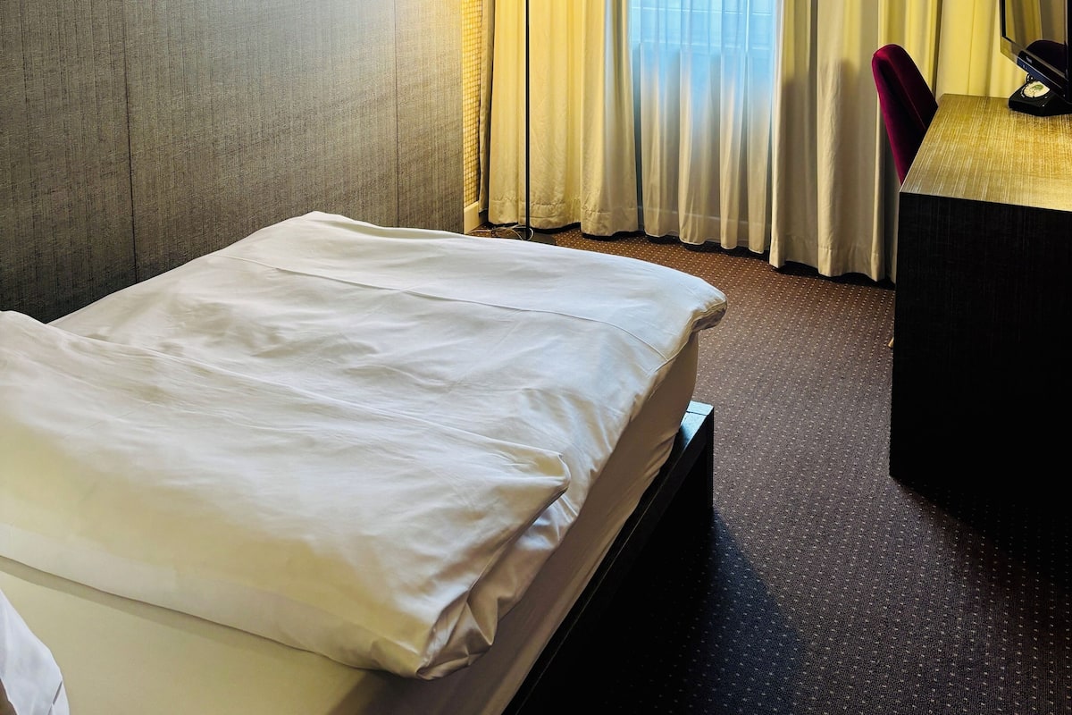 Une chambre d’hôtel avec un lit, un bureau avec un ordinateur, une lampe et une fenêtre avec des rideaux.