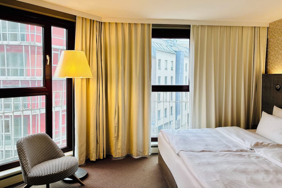 Une chambre d’hôtel avec un lit, une chaise, une lampe et de grandes fenêtres équipées de rideaux.