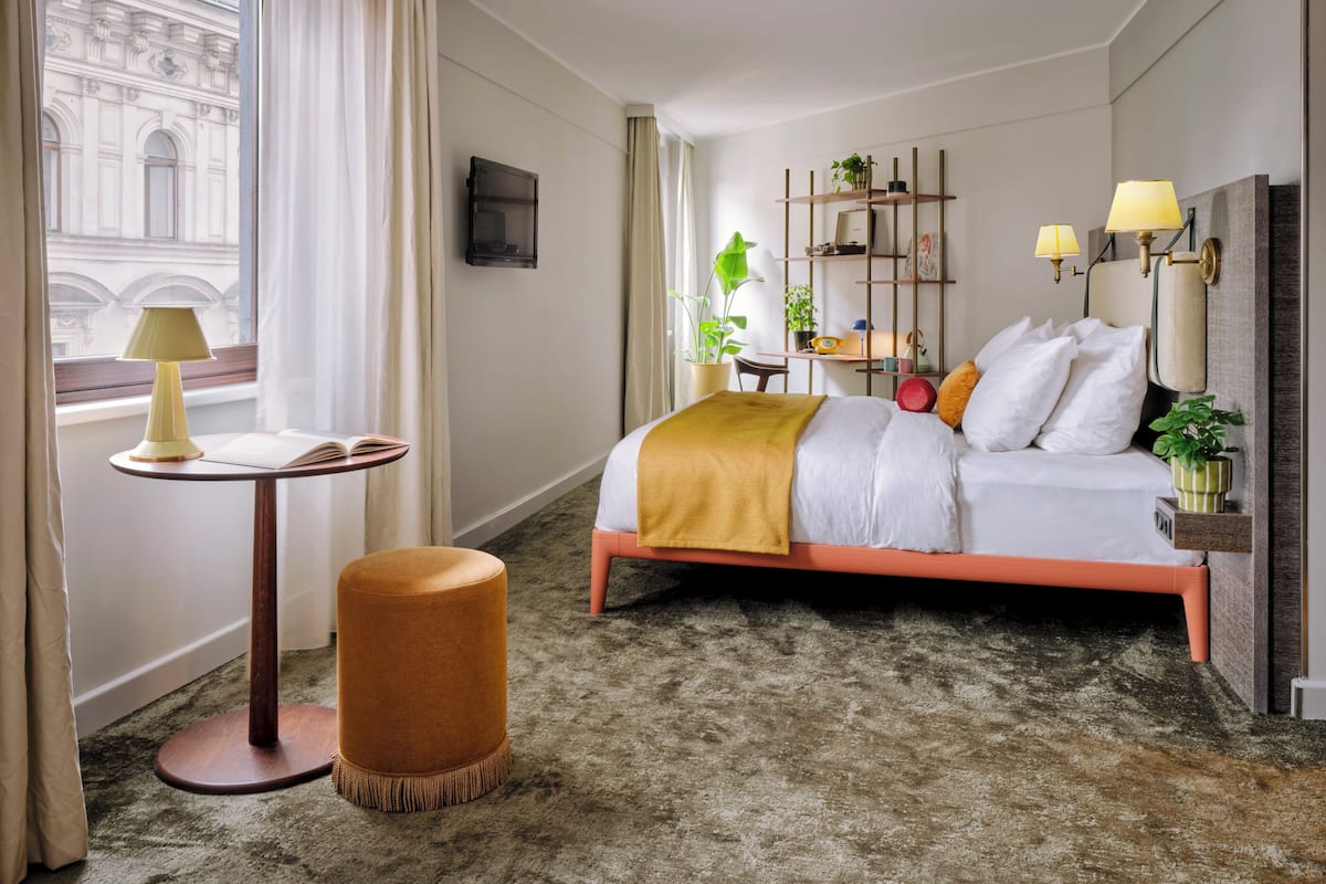 Une chambre d’hôtel comprenant un lit, une petite table avec une lampe, un pouf rond, un bureau avec une chaise et une bibliothèque.