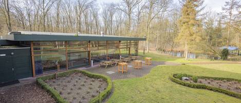 A modern building with large glass windows and a patio area with wooden tables and chairs.