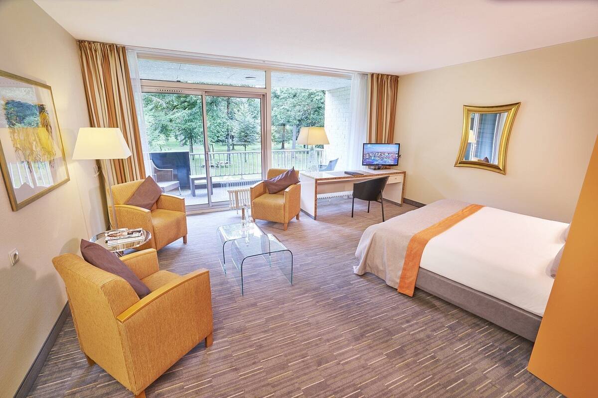 A hotel room with a bed, two armchairs, a desk, a television, and a balcony with a view of trees.