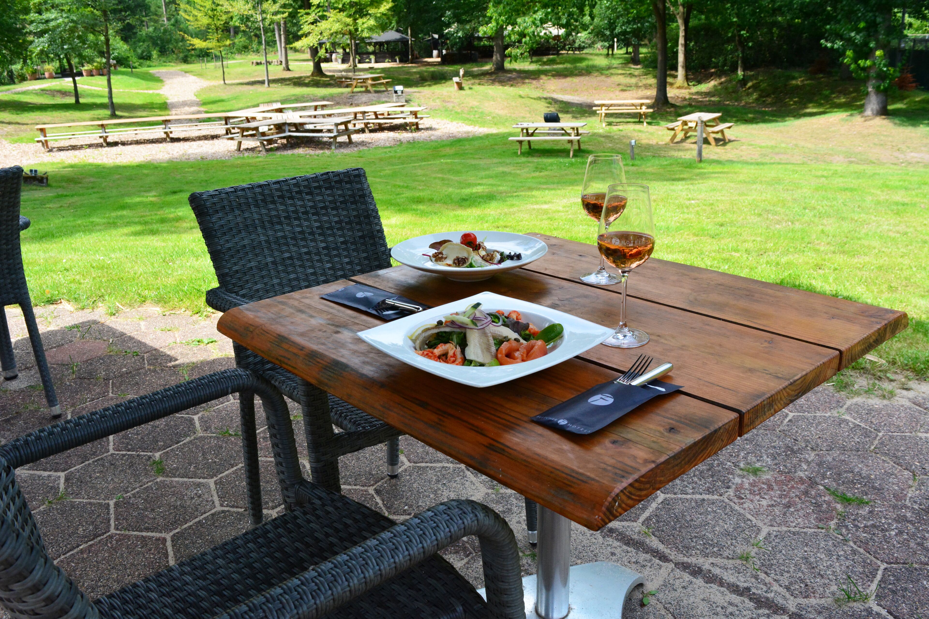 Un área de comedor al aire libre con una mesa de madera, sillas de mimbre y vistas a un parque con mesas de picnic.