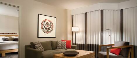 A modern hotel room with a sofa, a wooden coffee table, a chair, a bed, and a framed artwork on the wall.