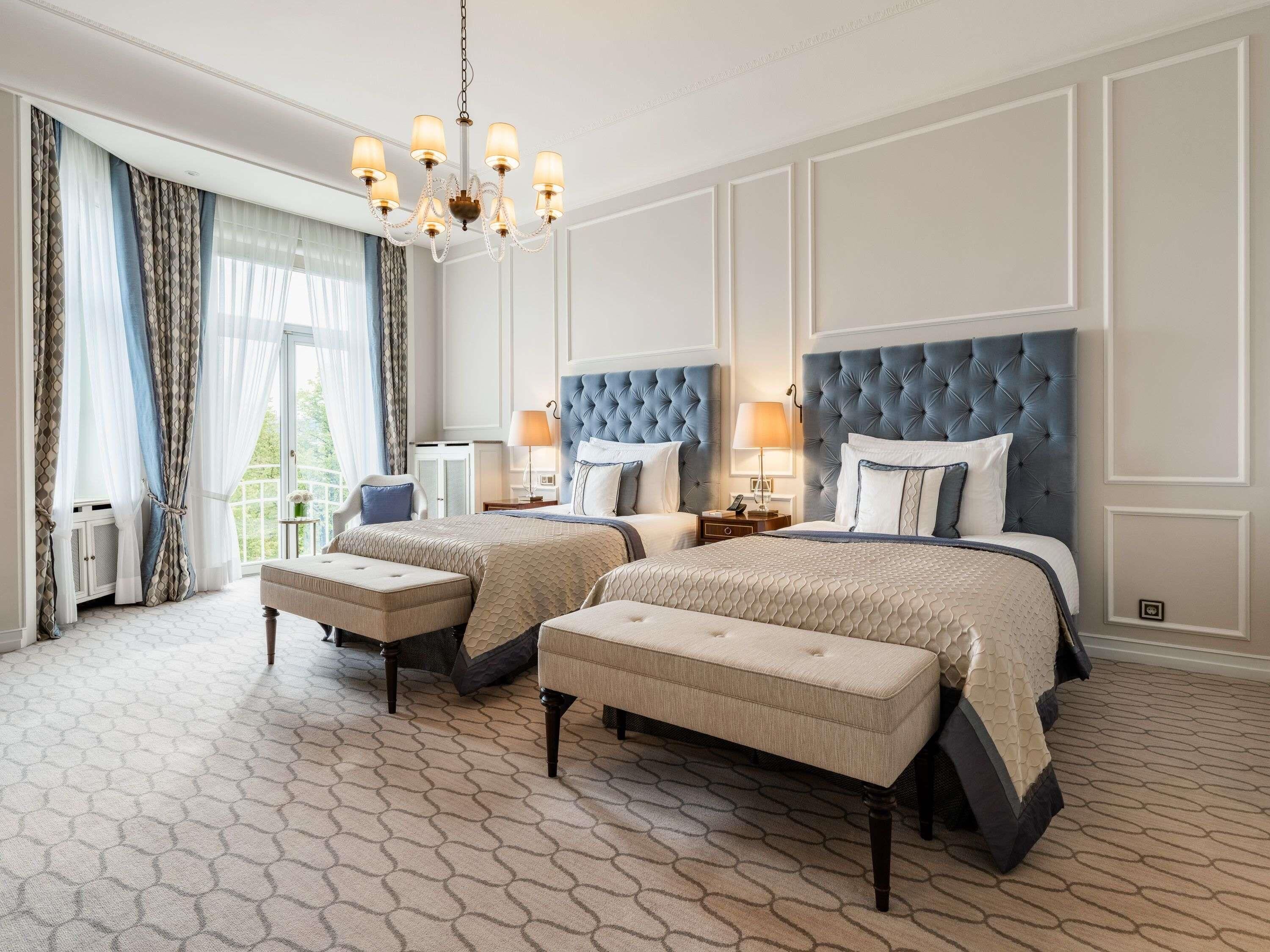 Quarto de hotel com duas camas, um candelabro e vista para o verde lá fora.