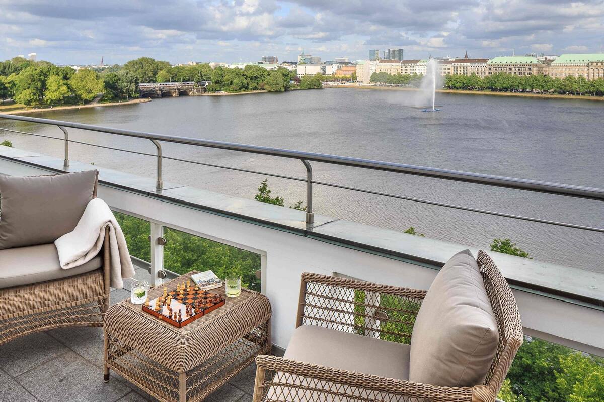 A balcony with wicker furniture, a chessboard, and a view of a river with a fountain.