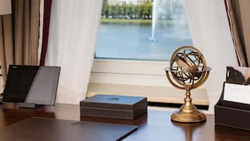 Un escritorio con una computadora portátil, un globo terráqueo y una caja, cerca de una ventana con cortinas y vistas a una fuente y edificios.