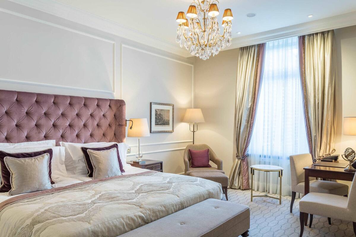A hotel room with a tufted headboard, a large bed with multiple pillows, a bedside table with a lamp, a chair, and a small table with a globe.