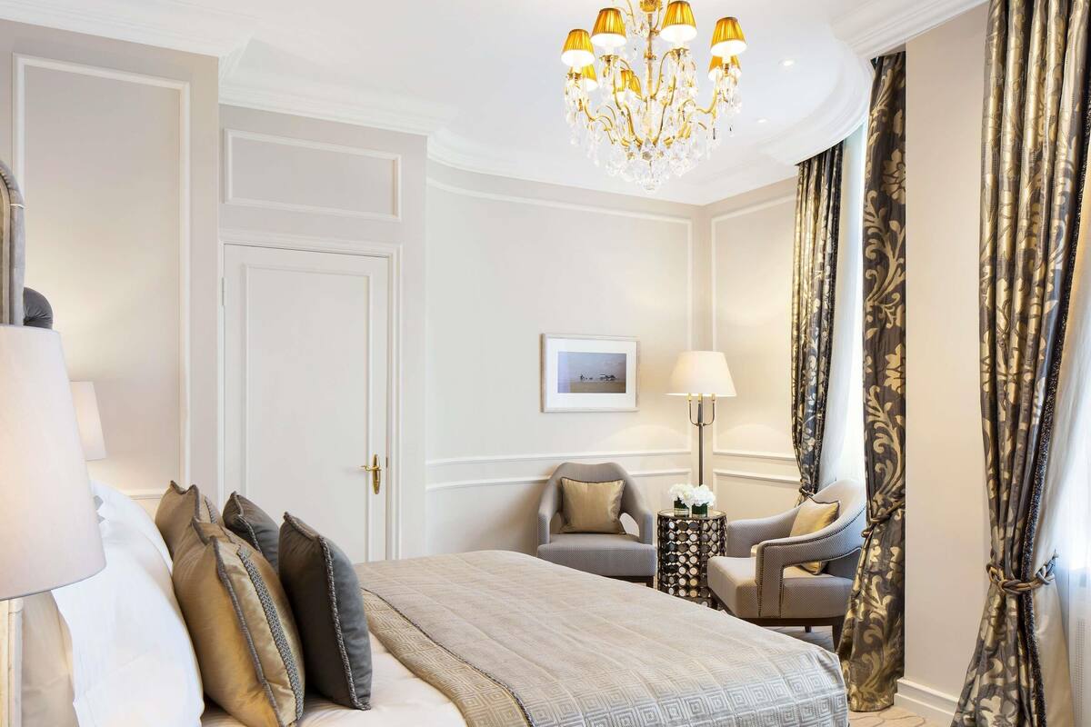 A hotel room with a large bed, a chandelier, a side table with a lamp, and a view of the outside through curtains.