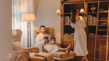 A man reading in a chair, a woman standing by a bookshelf, and a lamp on a side table.