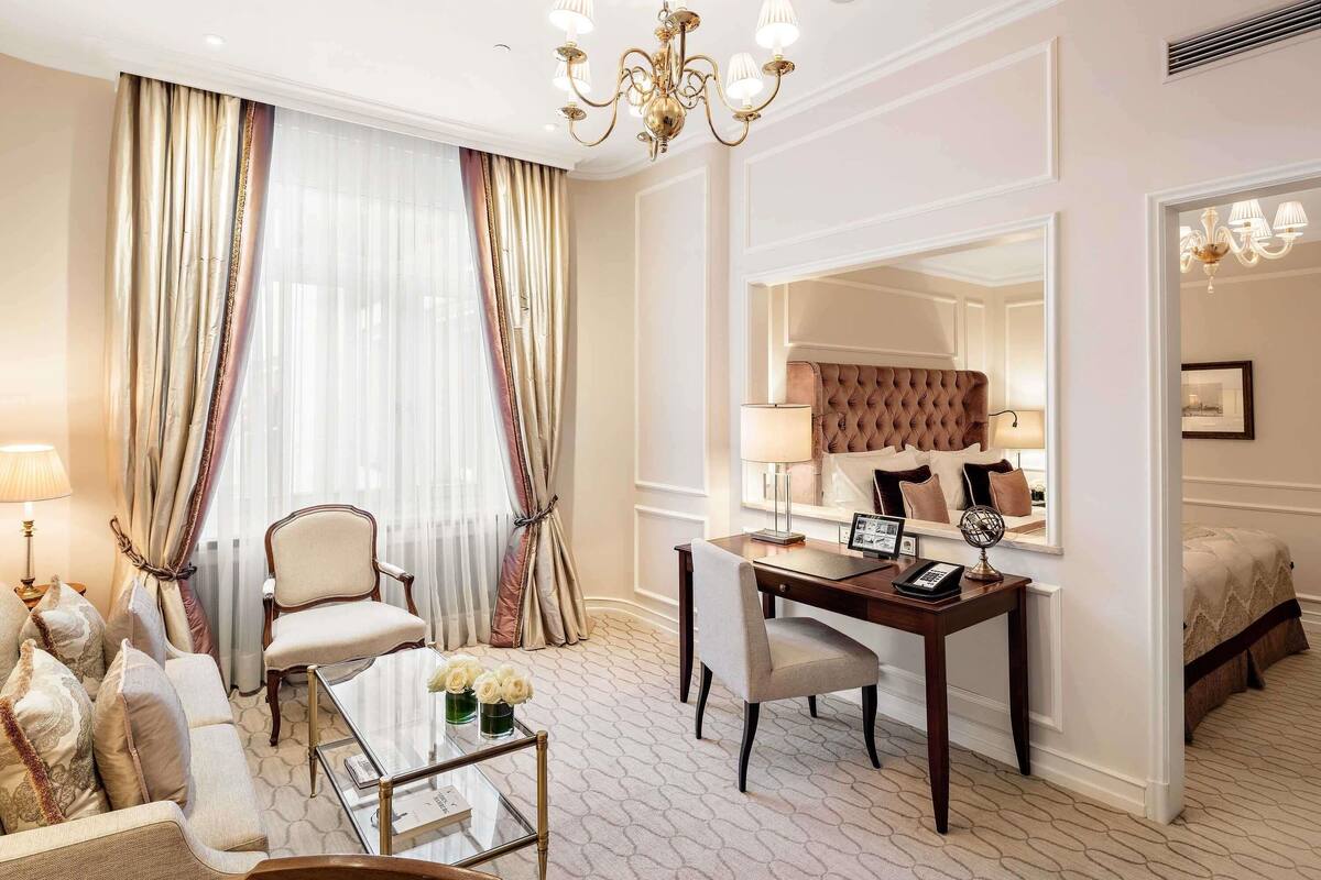 A hotel room with a bed, a desk with a chair, a sofa, a glass coffee table, and a chandelier.