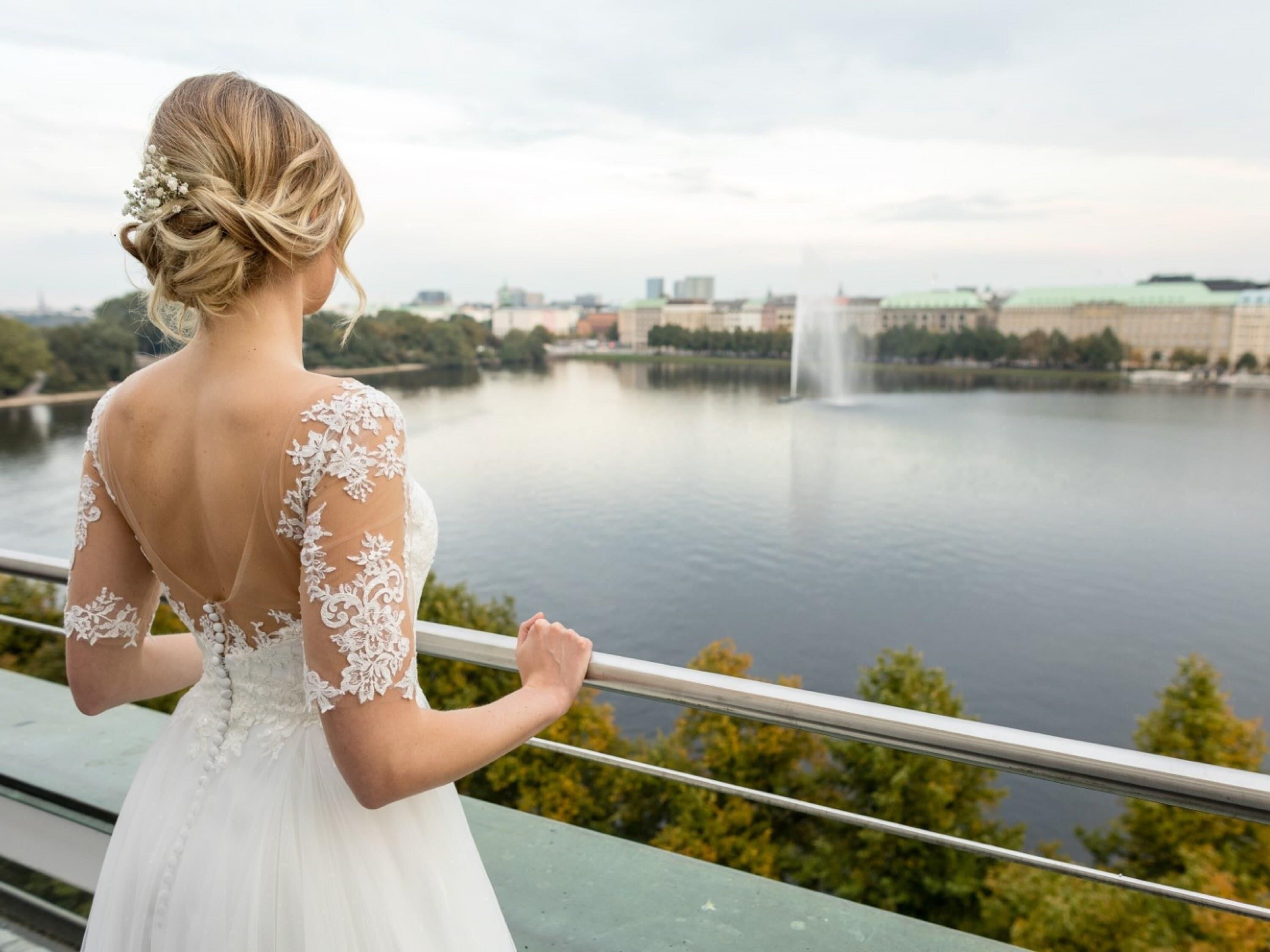 Eine Person in einem weißen Brautkleid mit Spitzenbesatz steht auf einem Balkon und blickt auf ein Gewässer mit einem Springbrunnen und Gebäuden im Hintergrund.