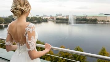 Eine Person in einem weißen Brautkleid mit Spitzenbesatz steht auf einem Balkon und blickt auf ein Gewässer mit einem Springbrunnen und Gebäuden im Hintergrund.