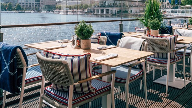 A waterfront restaurant with outdoor seating, a view of the river, and city buildings in the background. - Fairmont Hotel Vier Jahreszeiten Hamburg (Hamburg)