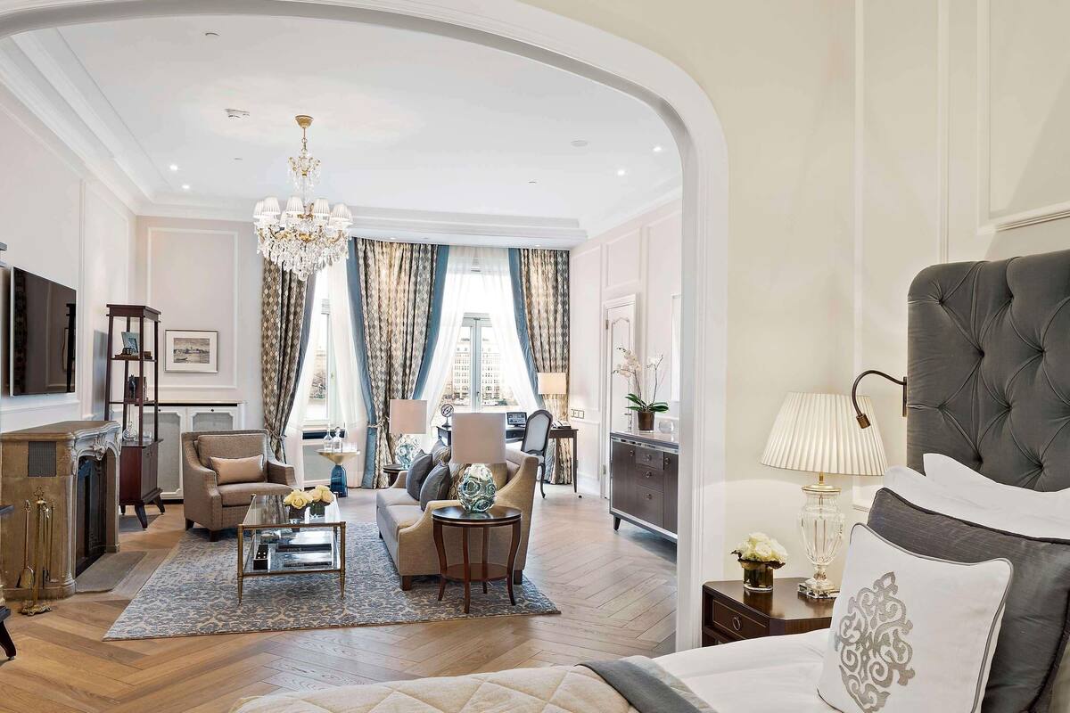 A spacious hotel room with a large bed, a sitting area with a sofa and chairs, a television, and a view of the city through large windows.