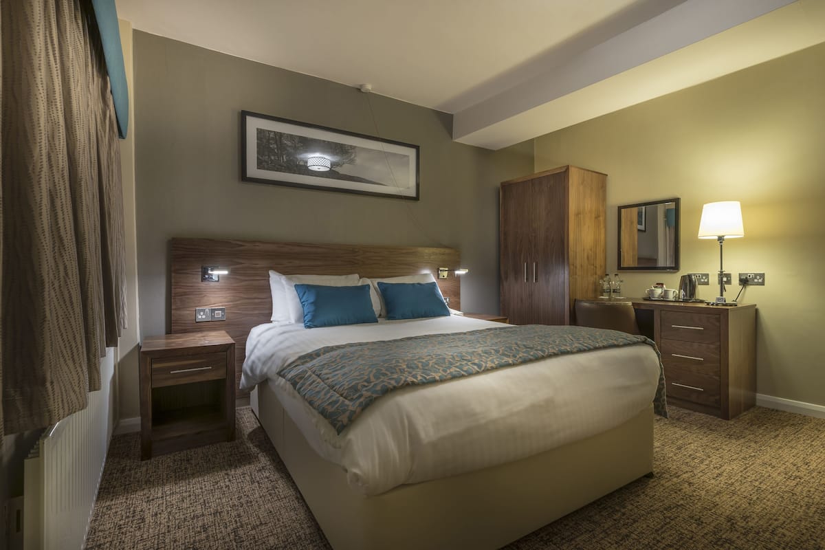 A hotel room with a large bed, a wooden headboard, a nightstand, a dresser, a lamp, and a framed picture on the wall.