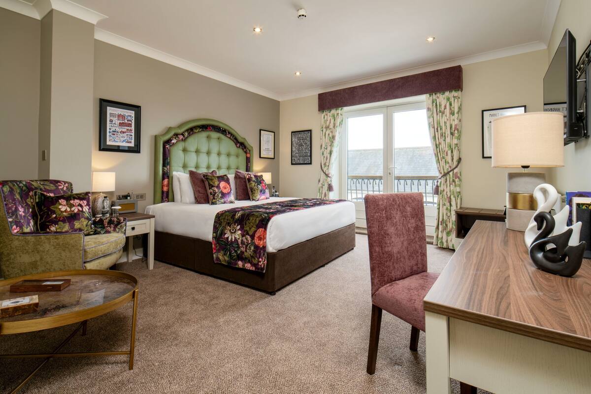 A hotel room with a bed, a desk, a chair, a nightstand, a lamp, and a window with curtains.