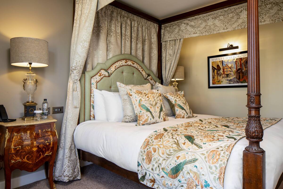 A four-poster bed with a patterned bedspread, a nightstand with a lamp, and a framed picture on the wall.