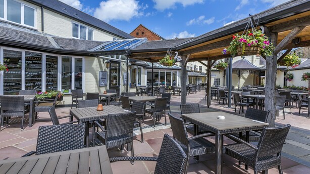 An outdoor patio area with tables and chairs, a hanging flower basket, and a building with large windows in the background. - The Three Swans Hotel, Market Harborough, Leicestershire (Market Harborough)