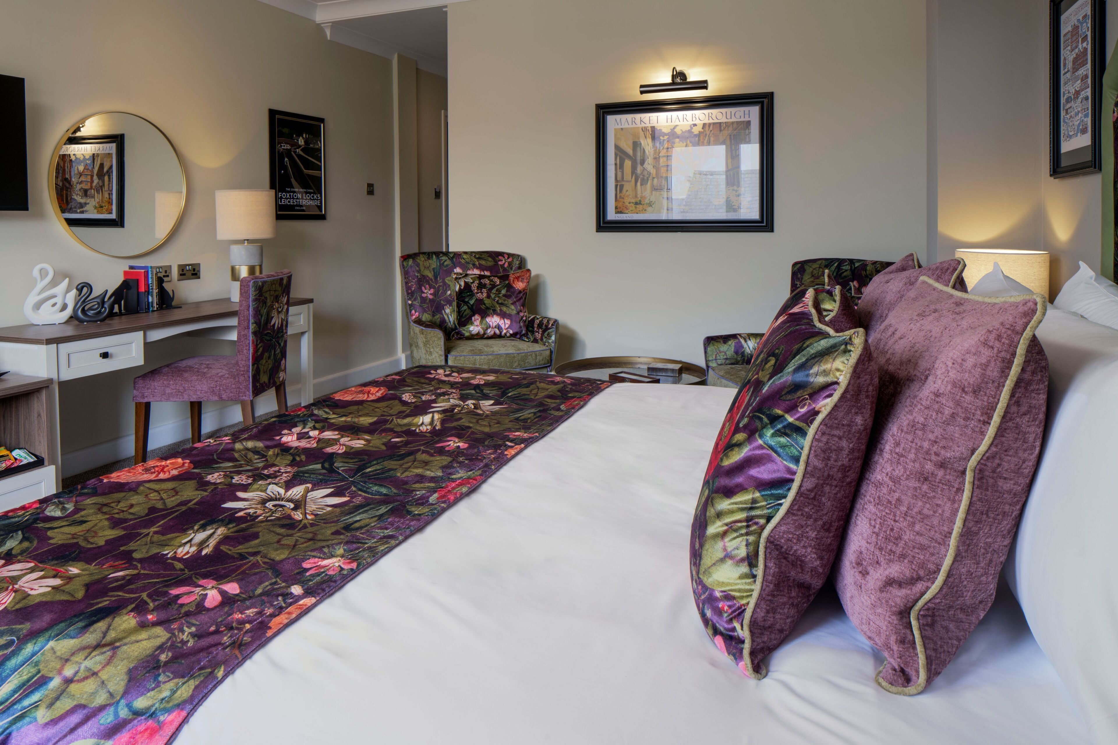 A hotel room with a large bed, a desk, two chairs, a round mirror, and framed pictures on the walls.