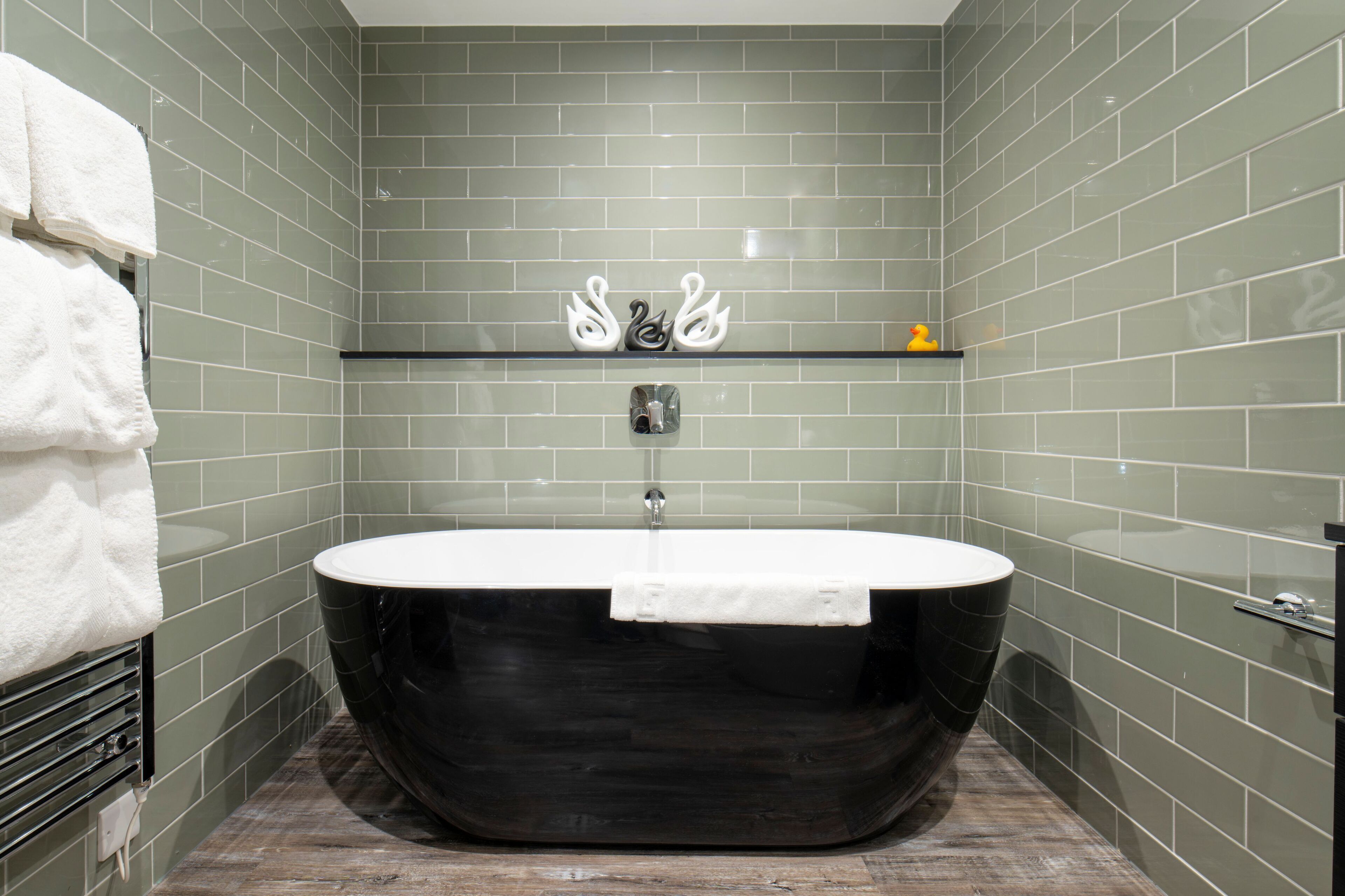 A modern bathroom with a freestanding bathtub, green tiled walls, a towel rack, and a small shelf with a rubber duck.