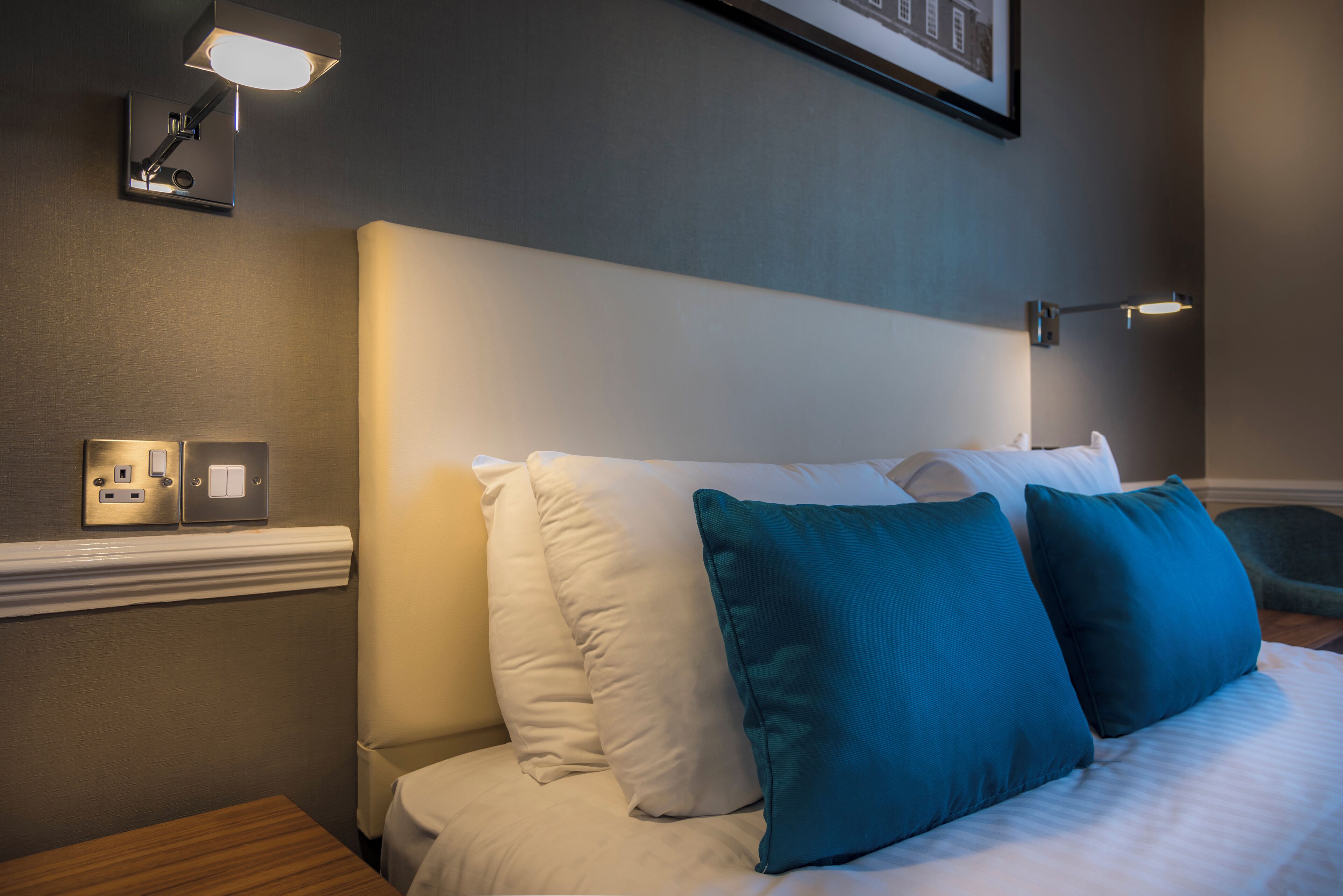 Un lit bien fait, agrémenté d’oreillers blancs et turquoise, un luminaire fixé au mur et un tableau encadré au-dessus du lit.
