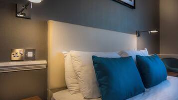 Un lit bien fait, agrémenté d’oreillers blancs et turquoise, un luminaire fixé au mur et un tableau encadré au-dessus du lit.