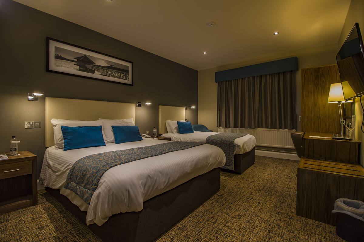 A hotel room with two beds, a large window with curtains, a bedside table with a lamp, and a framed picture on the wall.