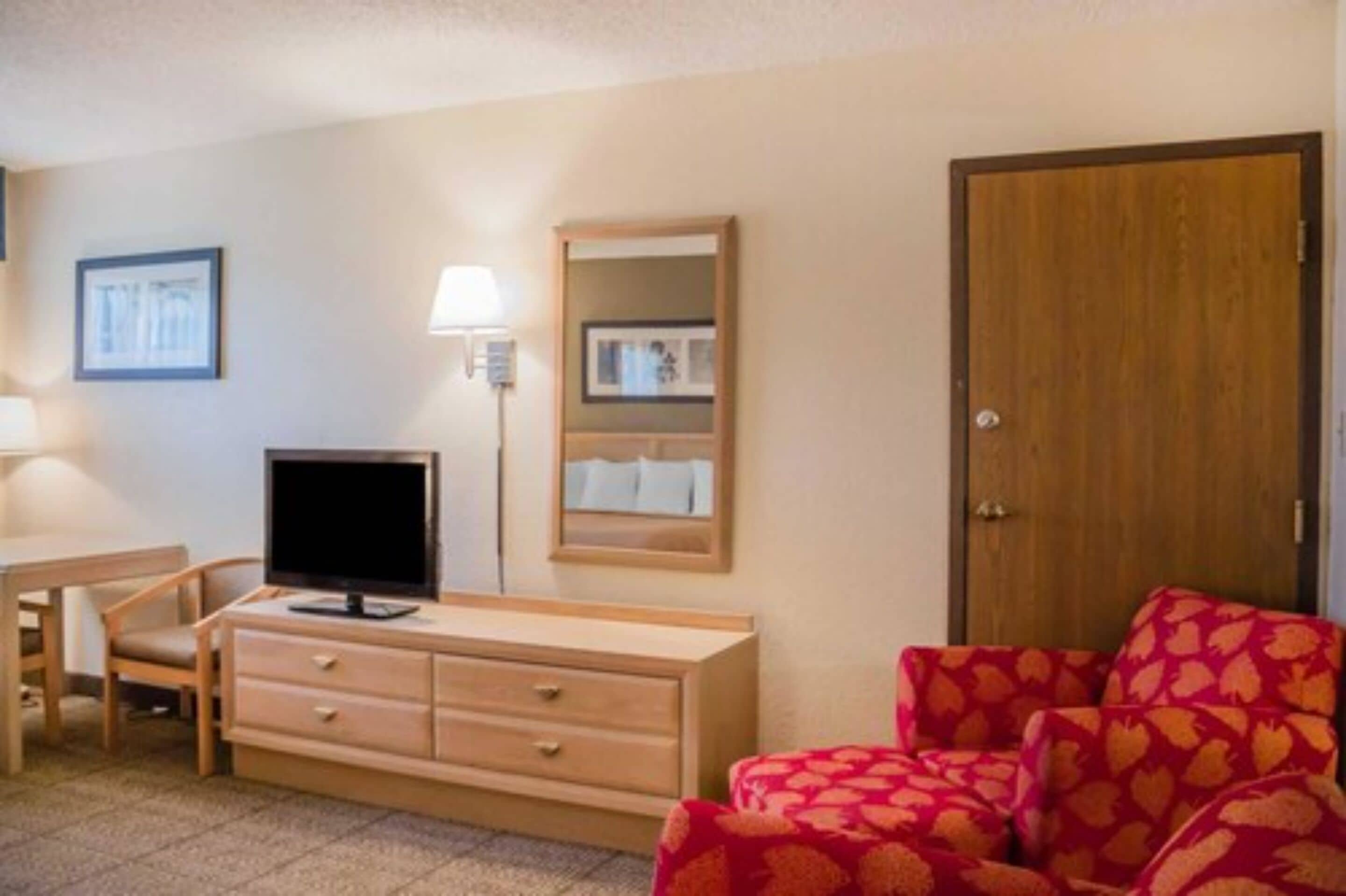 A hotel room with a wooden dresser, a television, a red patterned sofa, a wooden door, and framed pictures on the wall.