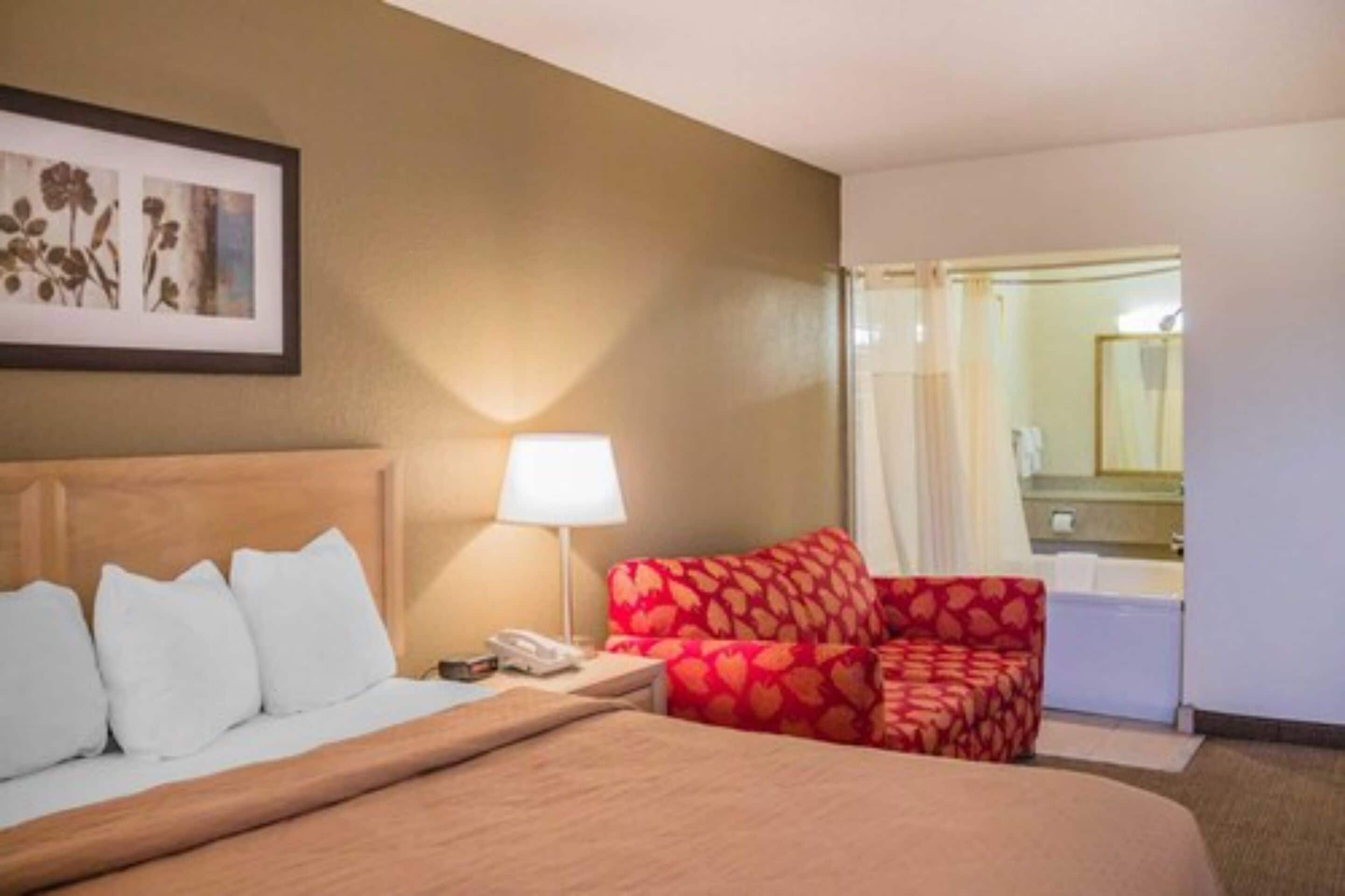Habitación de hotel con cama, mesita de noche, sofá rojo estampado, lámpara y un cuadro en la pared.