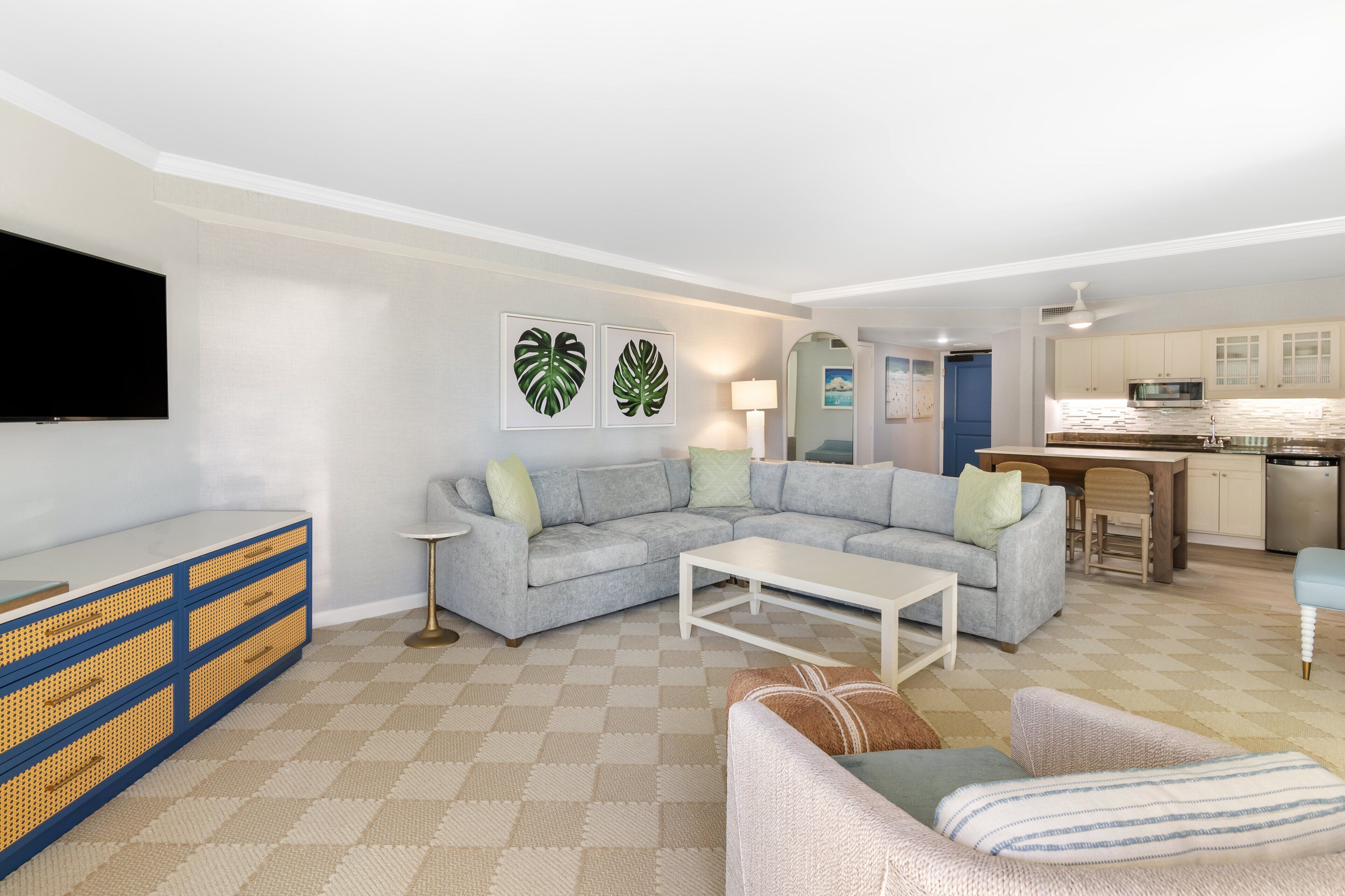 A living room with a grey sofa, a white coffee table, and a TV on a blue and yellow cabinet. There is a kitchen in the background with a blue door.