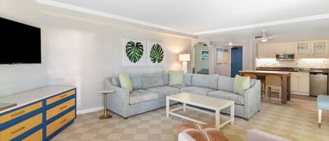 A living room with a grey sofa, a white coffee table, and a TV on a blue and yellow cabinet. There is a kitchen in the background with a blue door.