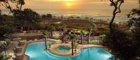 A pool area with a view of the ocean at sunset, surrounded by palm trees and lounge chairs.